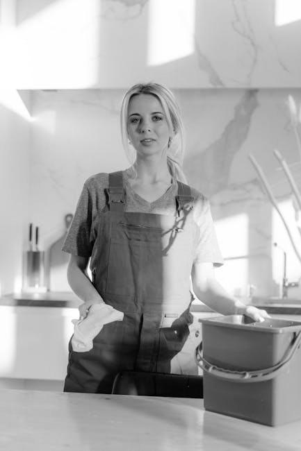 A young woman with blonde hair, dressed in a casual grey T-shirt and dark overalls, stands in a modern kitchen. She is holding a cleaning cloth in her right hand and resting her left hand on a plastic